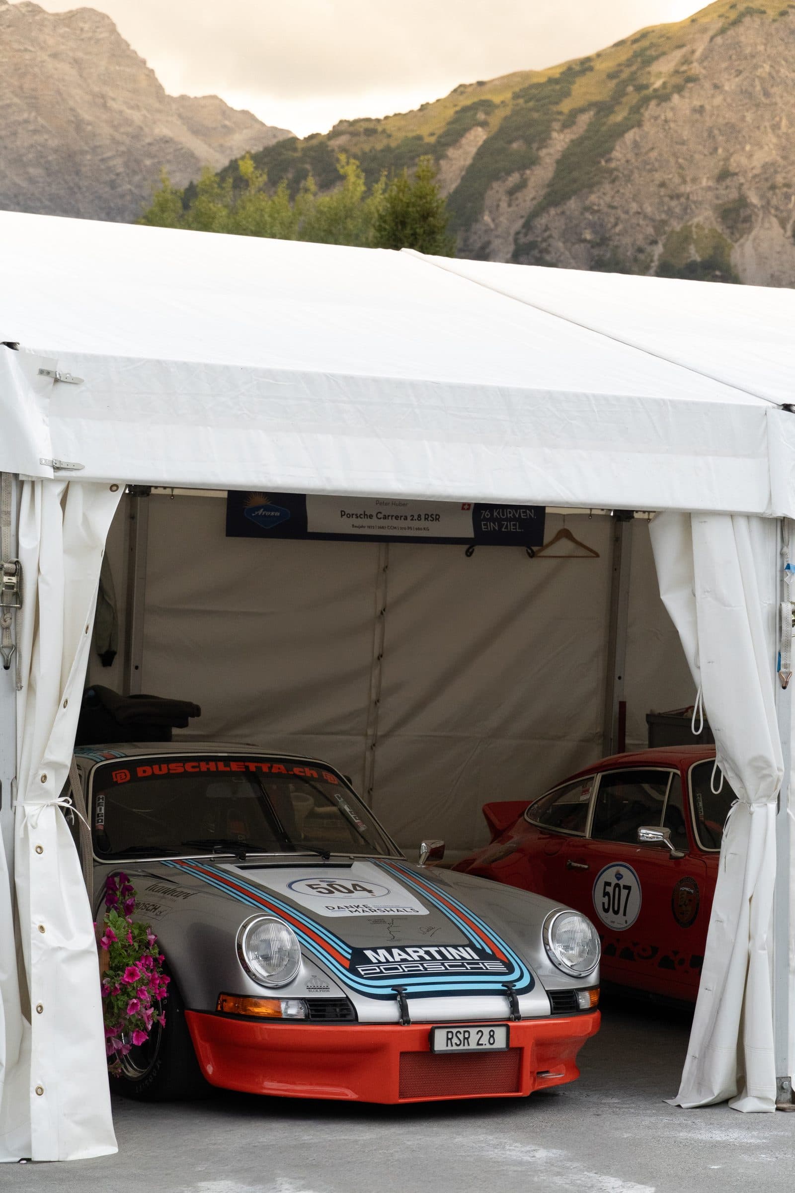 Porsche 911 in Martini-Lackierung im Fahrerlager, Bergpanorama im Hintergrund